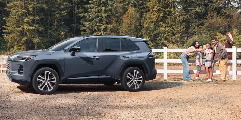 A gray Toyota RAV4 parked beside a family taking a photo by a white fence with trees in the background.